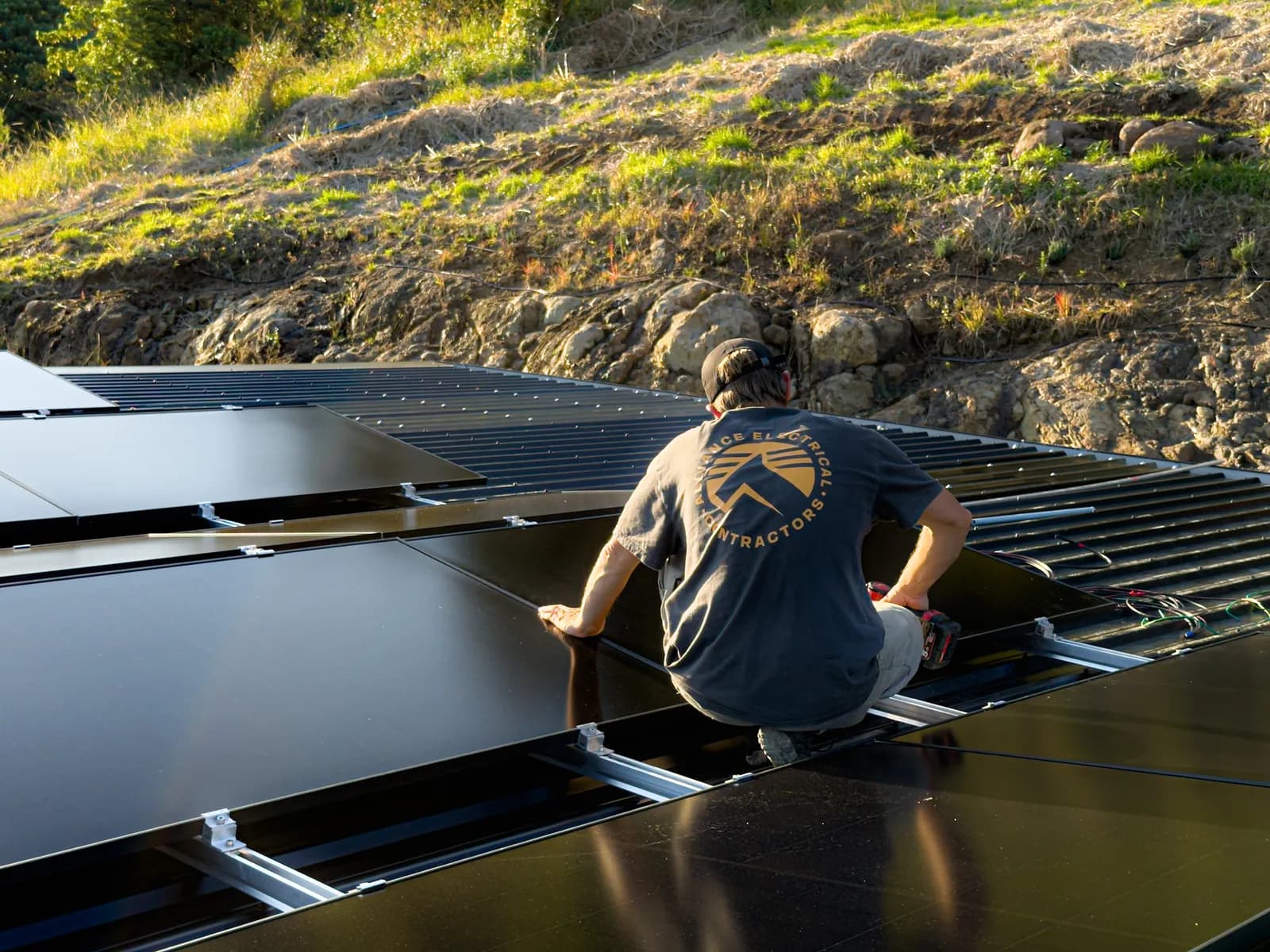An Alliance Electrical electrician in a branded work shirt fitting solar panels to a roof in late afternoon light, with a green hillside behind.
